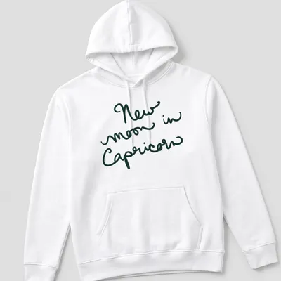 Handwritten script of 'New moon in Capricorn' in dark green text — printed on a unisex hoodie