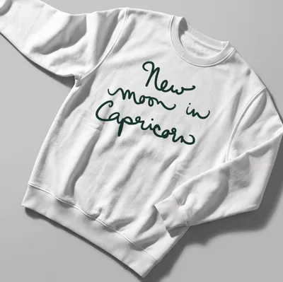 Handwritten script of 'New moon in Capricorn' in dark green text — printed on a unisex sweatshirt