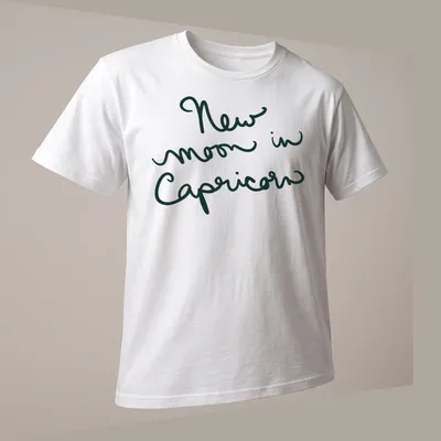 Handwritten script of 'New moon in Capricorn' in dark green text — printed on a unisex t-shirt (angled view)