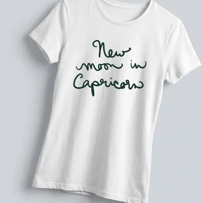 Handwritten script of 'New moon in Capricorn' in dark green text — printed on a women's shirt